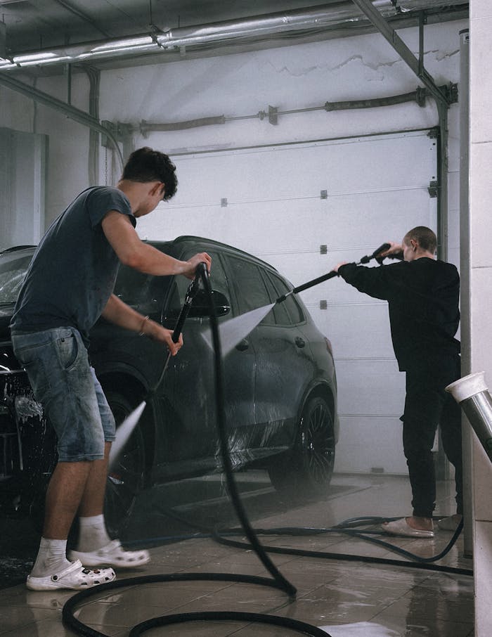 Two men using hoses to clean a black car in an indoor garage.